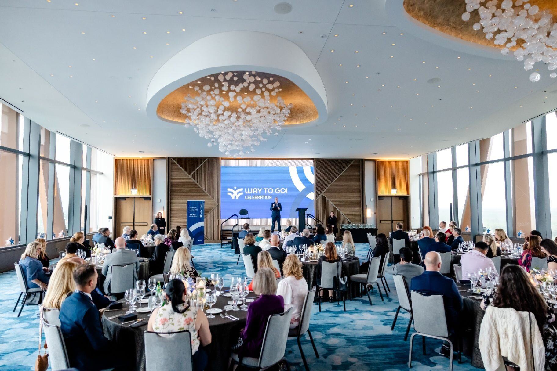 Full-room view of guests seated at dinner while a speaker presents at the front of a corporate celebration